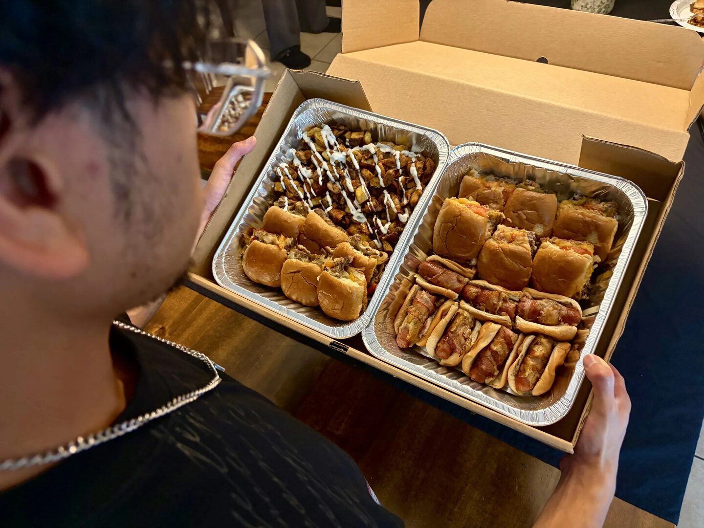 Two catering trays packed with sliders, bacon-wrapped hot dogs, and loaded potatoes.