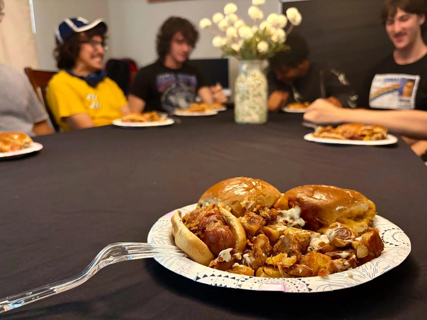 A plate of sliders, a bacon-wrapped hot dog, and loaded potatoes, with guests enjoying a meal around the table.