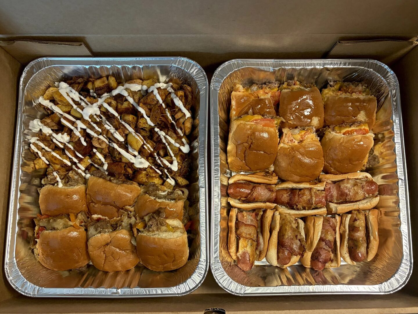 An overhead view of two catering trays filled with loaded potatoes, steak sliders, cheeseburger sliders, and bacon-wrapped hot dogs.